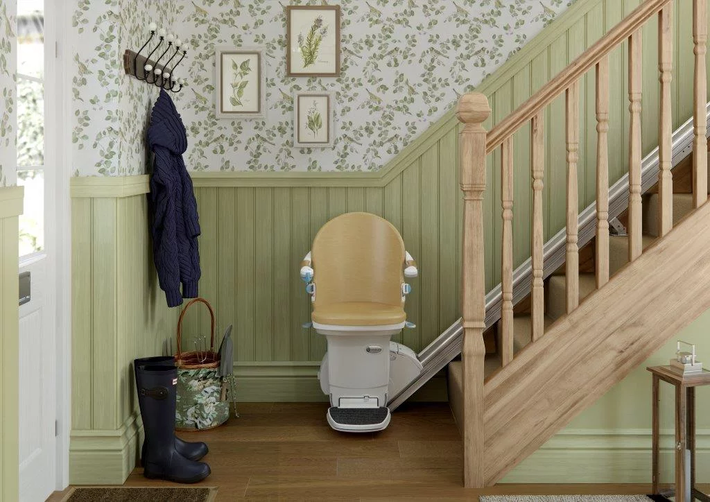 stairlift picture A stylish hallway featuring a wooden staircase with a beige stairlift at the bottom. The walls are decorated with green wainscoting and leafy wallpaper, with framed botanical prints. Nearby are a coat rack with a hanging sweater, a pair of black wellington boots, and a floral umbrella stand.