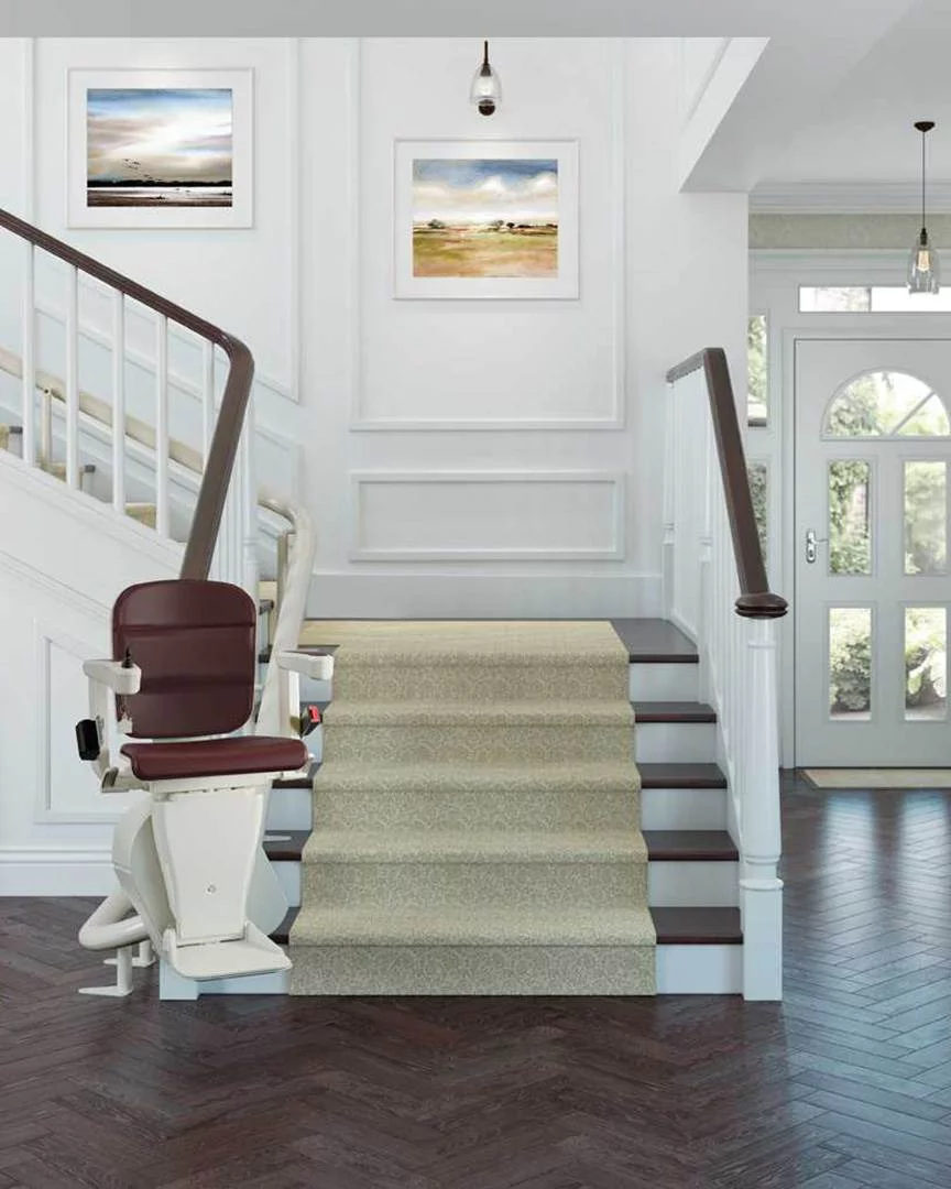 curved stairlift Elegant stairlift with a maroon cushioned seat and white base installed along a wide carpeted staircase in a stylish, well-lit hallway featuring modern artwork, white panelled walls, and dark herringbone flooring.