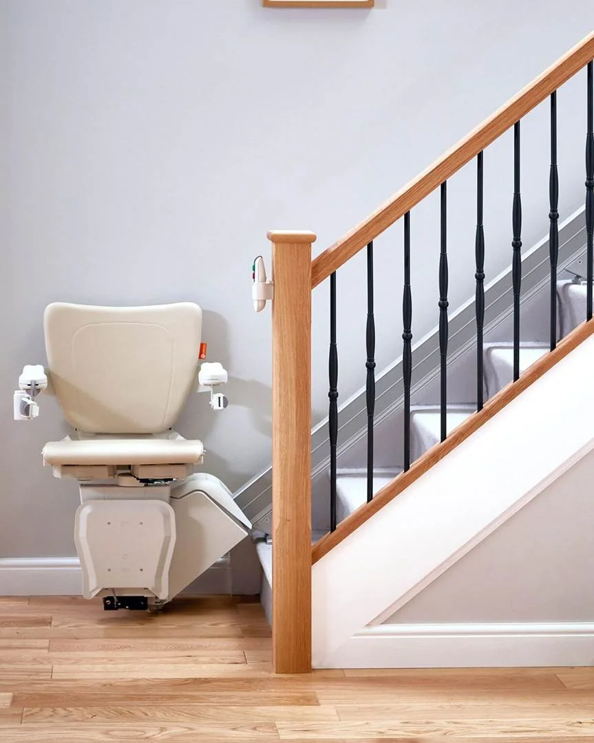 straight stairlift Modern indoor stairlift installed along a carpeted staircase with wooden handrail and black spindles, featuring a padded beige seat and foldable footrest positioned at the bottom of the stairs.
