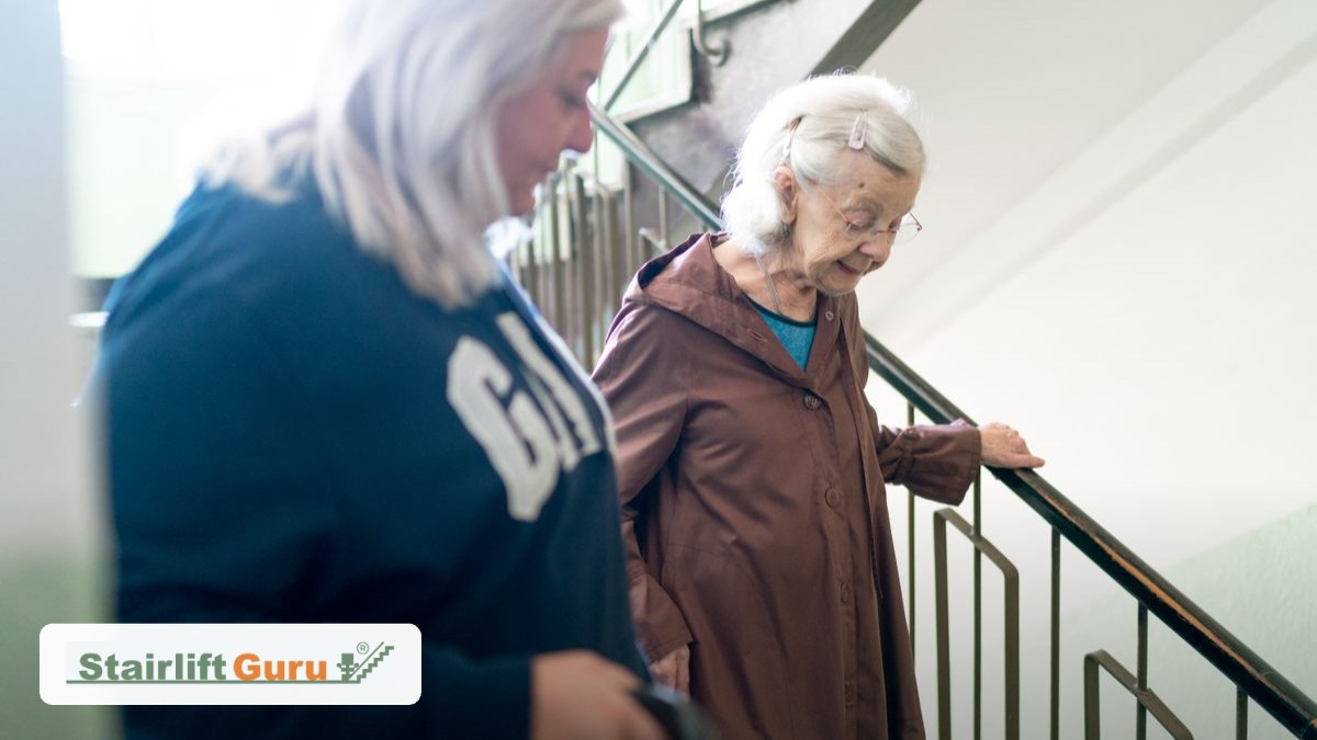 signs-elderly-parent-stairs-featured-v2 Two elderly women descend a staircase, one holding the railing while the other accompanies her; Stairlift Guru logo in the corner.
